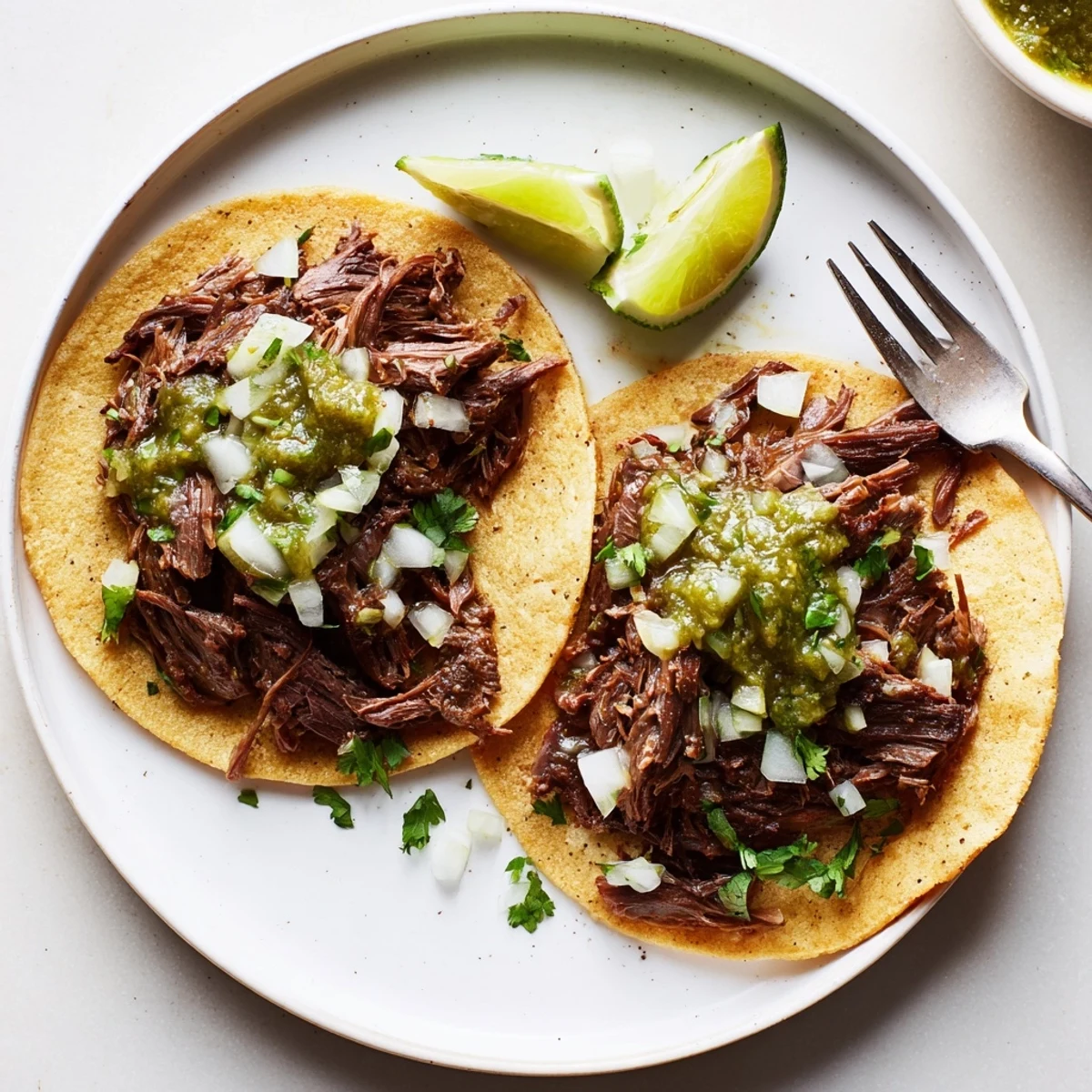 Close-up view of tender slow-braised beef cheek tacos topped with zesty salsa verde and lime wedges on a rustic serving plate. 