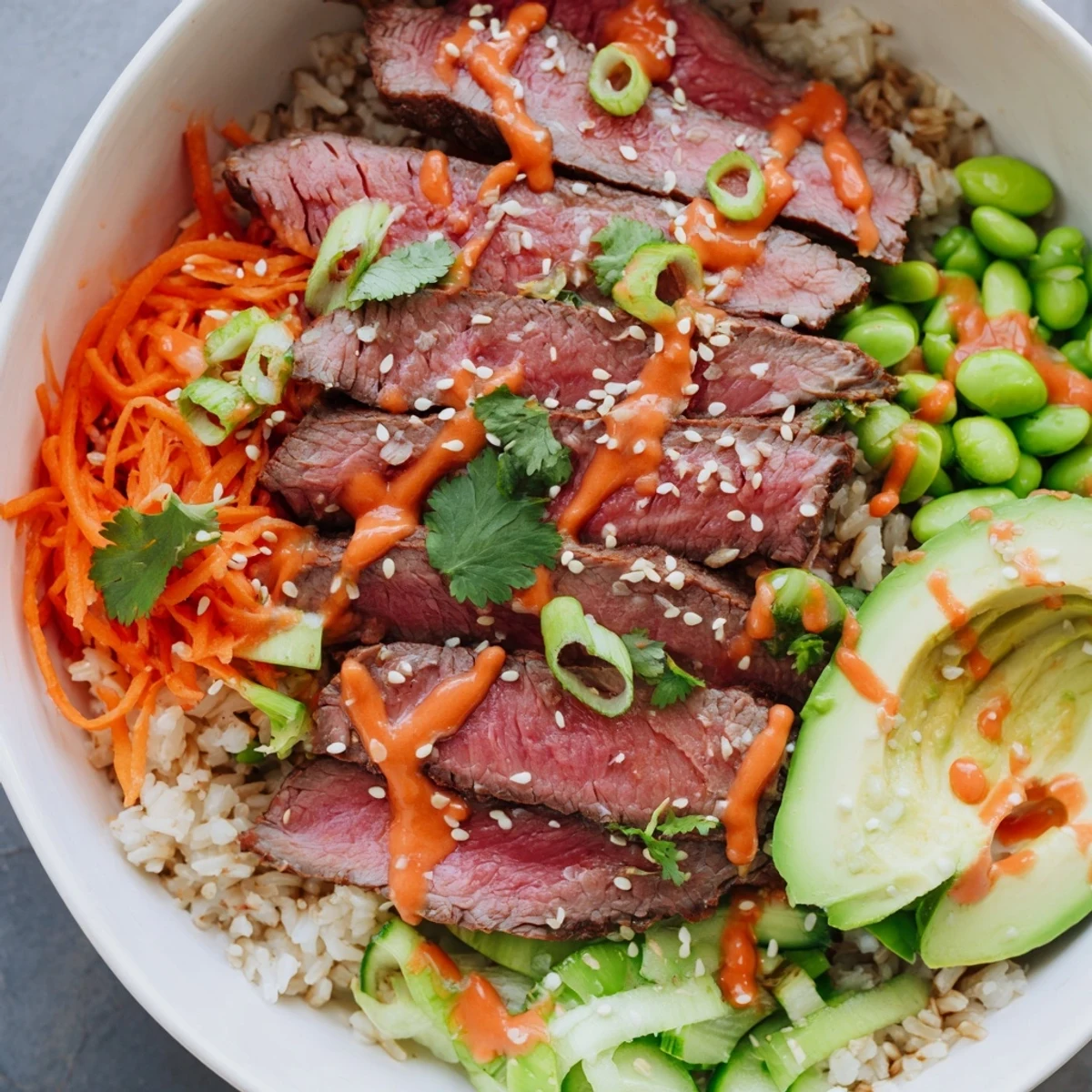 Colorful flank steak bowl with sliced beef, fresh vegetables, avocado, and spicy sriracha mayonnaise drizzled over steamed rice