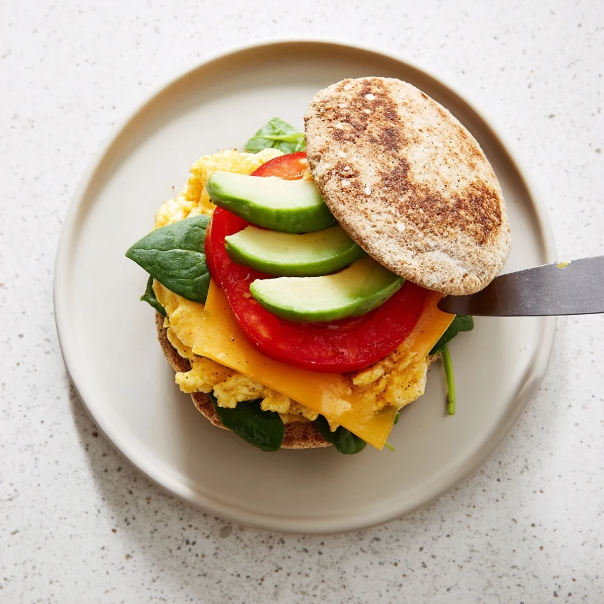 Nutritious healthy breakfast sandwich on a white plate showing egg, cheese, and fresh vegetable fillings spilling from toasted split English muffin