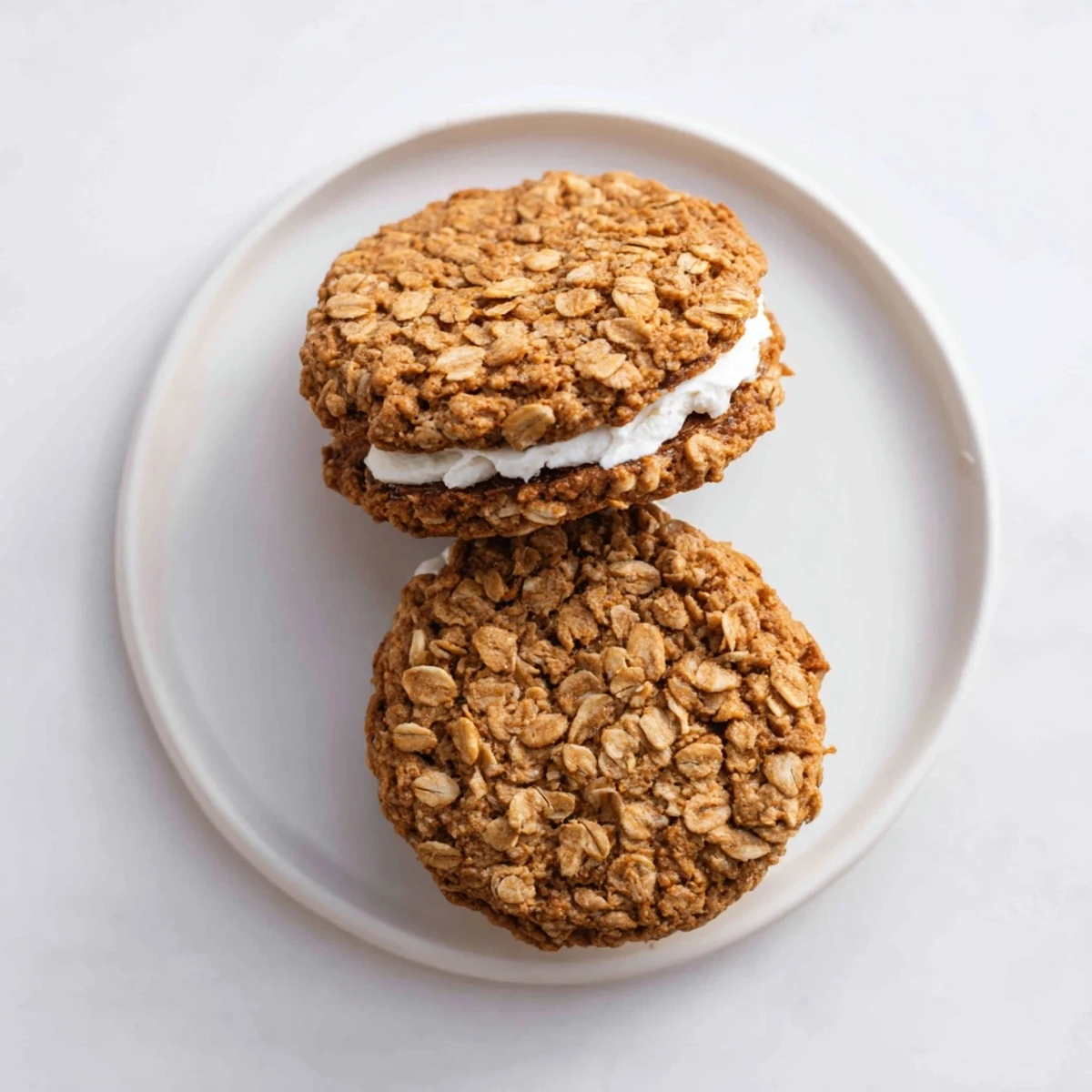 Two golden brown homemade oatmeal cream pies sandwiched with fluffy vanilla frosting on a white plate