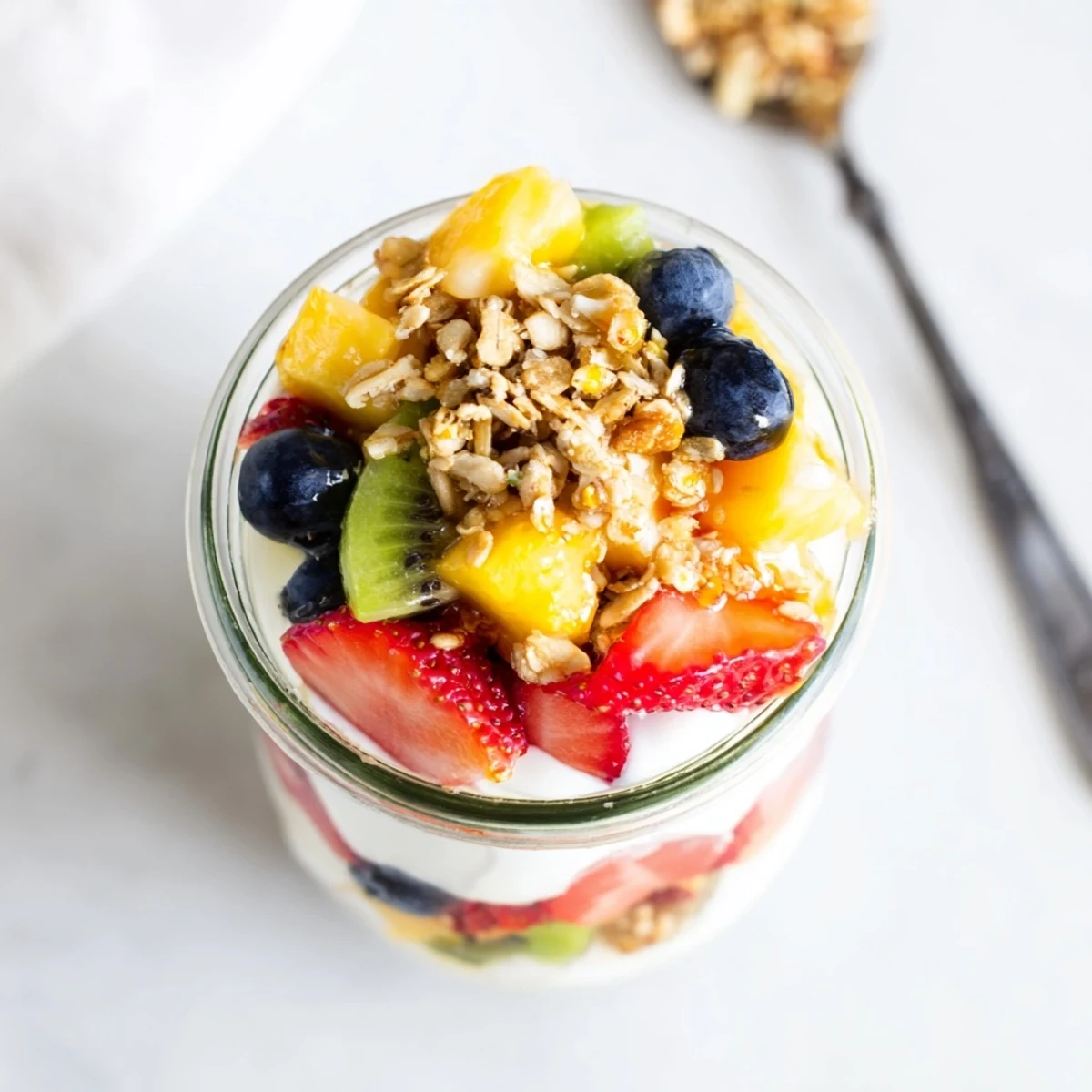 Glass jar displaying layered yogurt parfait with honey, strawberries, blueberries, mango, and kiwi slices topped with granola