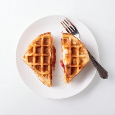 A close-up of Sweet Waffle Grilled Cheese on a white plate, sliced to reveal melted cheese and warm waffle texture.