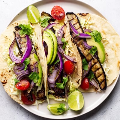 Golden-brown grilled eggplant on Charred Eggplant Tacos rests beside crisp lettuce, creamy avocado, and sliced red onion on a rustic plate.